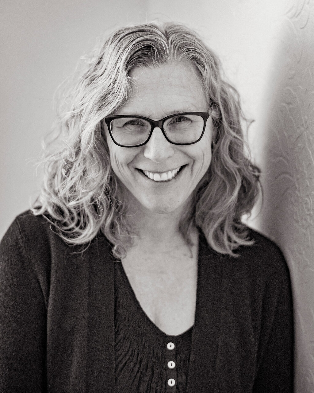 Photo Head shot of Laura MacGregor, she has shoulder length wavy hair. Wearing square lensed glasses and in a dark buttoned blouse, she is smiling widely.